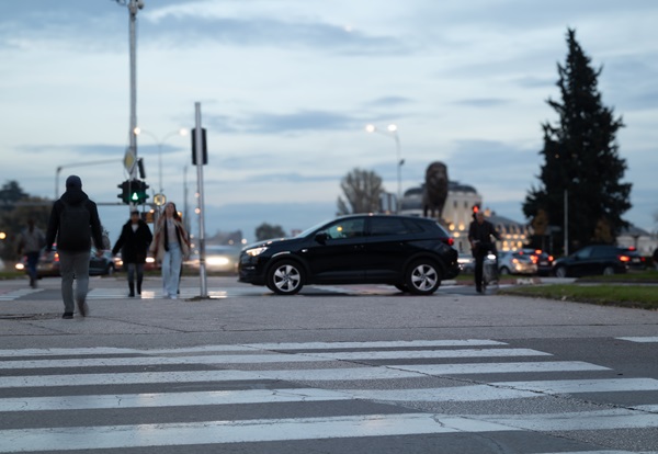 A black SUV stopped at a traffic light behind a white striped crosswalk as pedestrians and a bicyclist cross in the foreground and background at dusk.