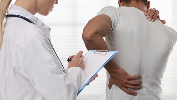 A medical professional in a white lab coat taking notes on a clipboard while a man in a white t-shirt stands with his back turned, clutching his painful neck and lower back.