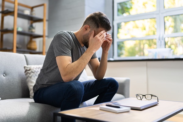 A man in a grey t-shirt sits on a sofa with his head in his hands, appearing to suffer from a persistent headache or post-concussion symptoms following a car accident in Delaware.